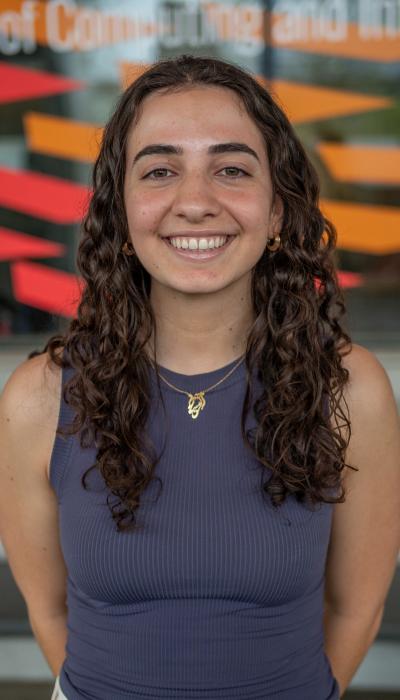 a female student with tan skin and long brown curly hair wearing a dark purple gray tank top poses in front of windows painted red and orange