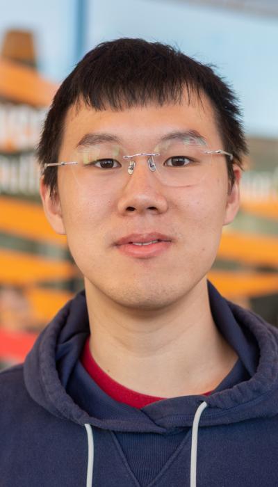 a male asian student with black hair, glasses and wearing a navy pullover hoodie stands in front of red and orange painted windows