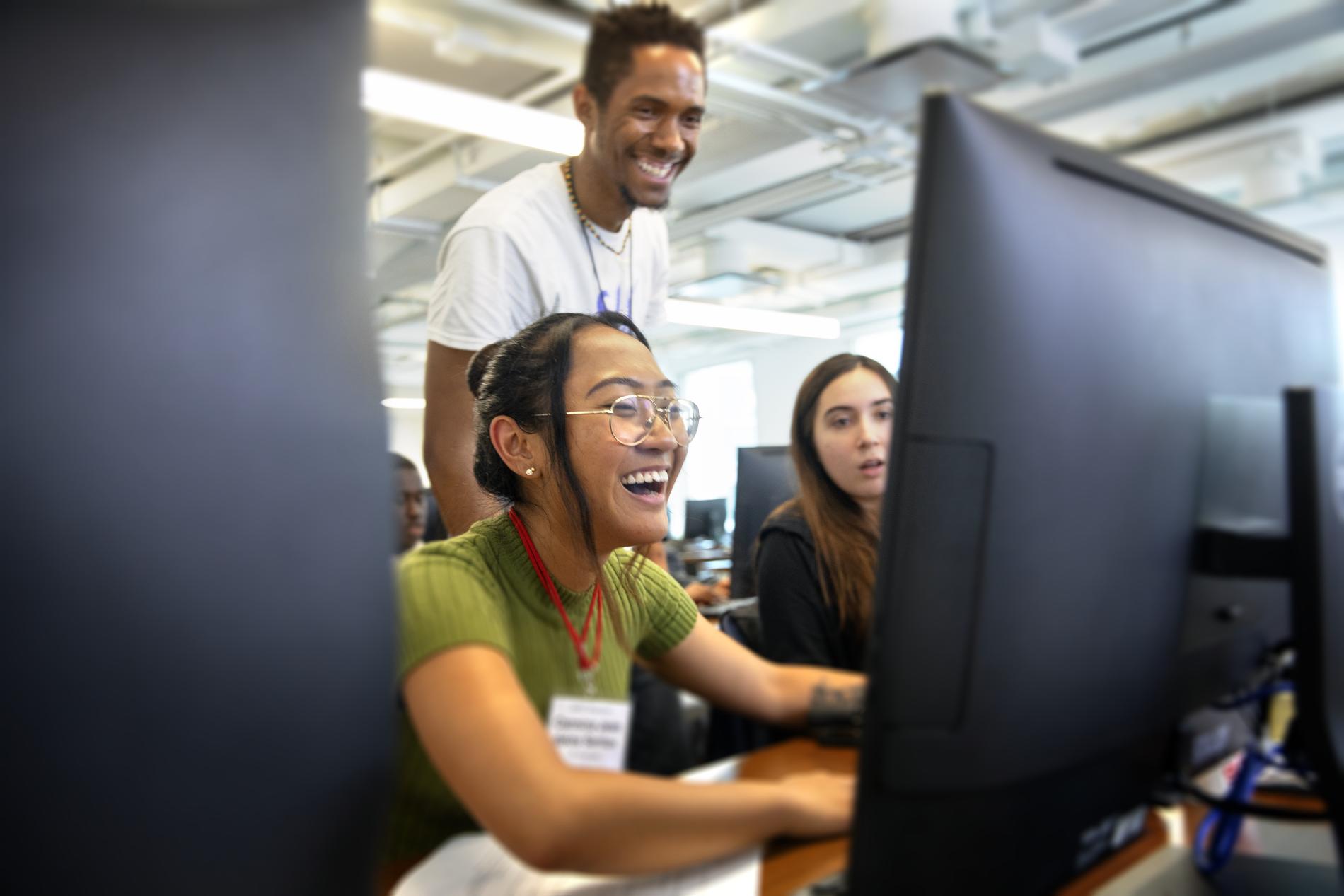 three students framed in the middle of two black computer desktops, smiling and working