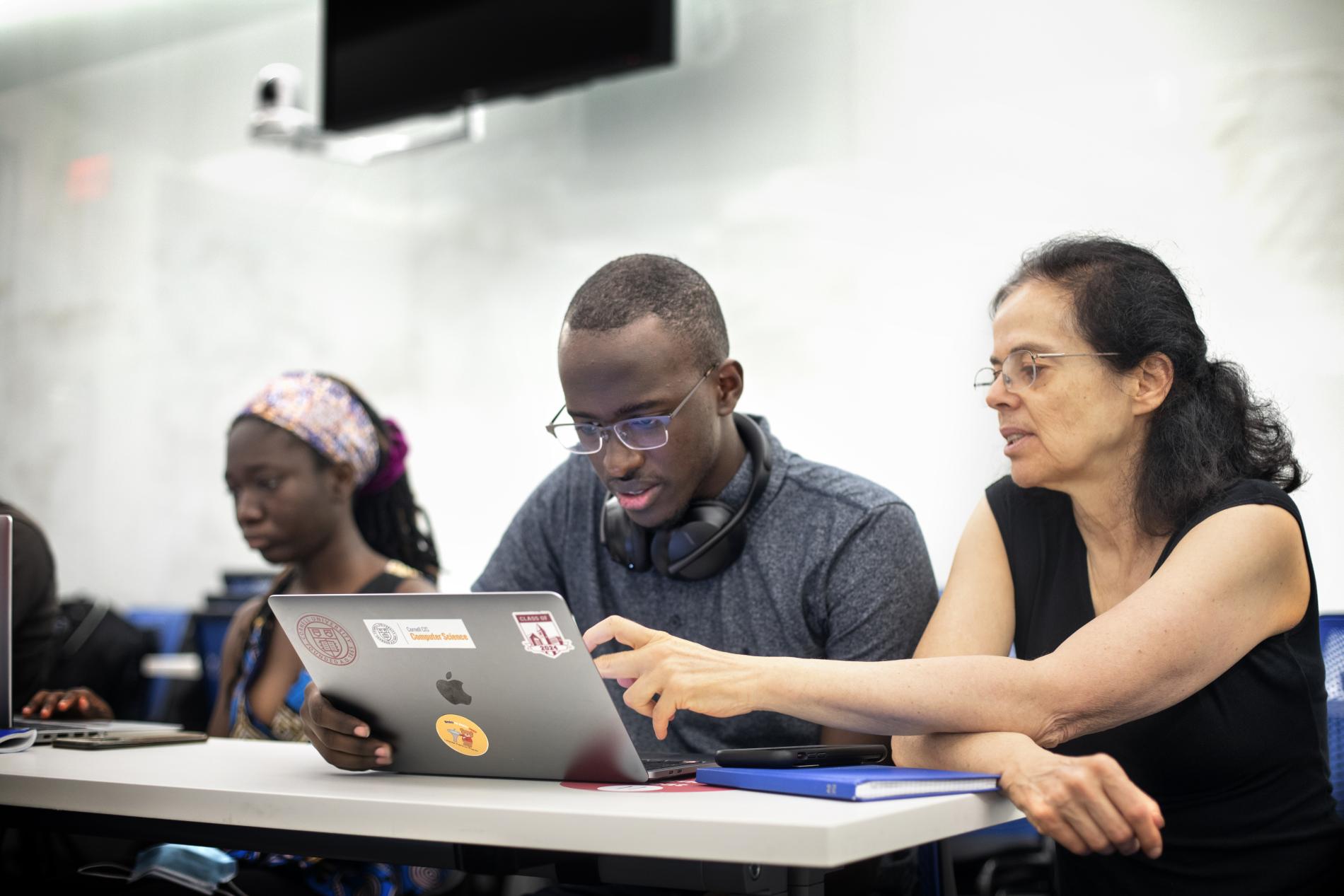 two black students sit at a table with the male student on a laptop working next to a woman faculty member pointing to the screen